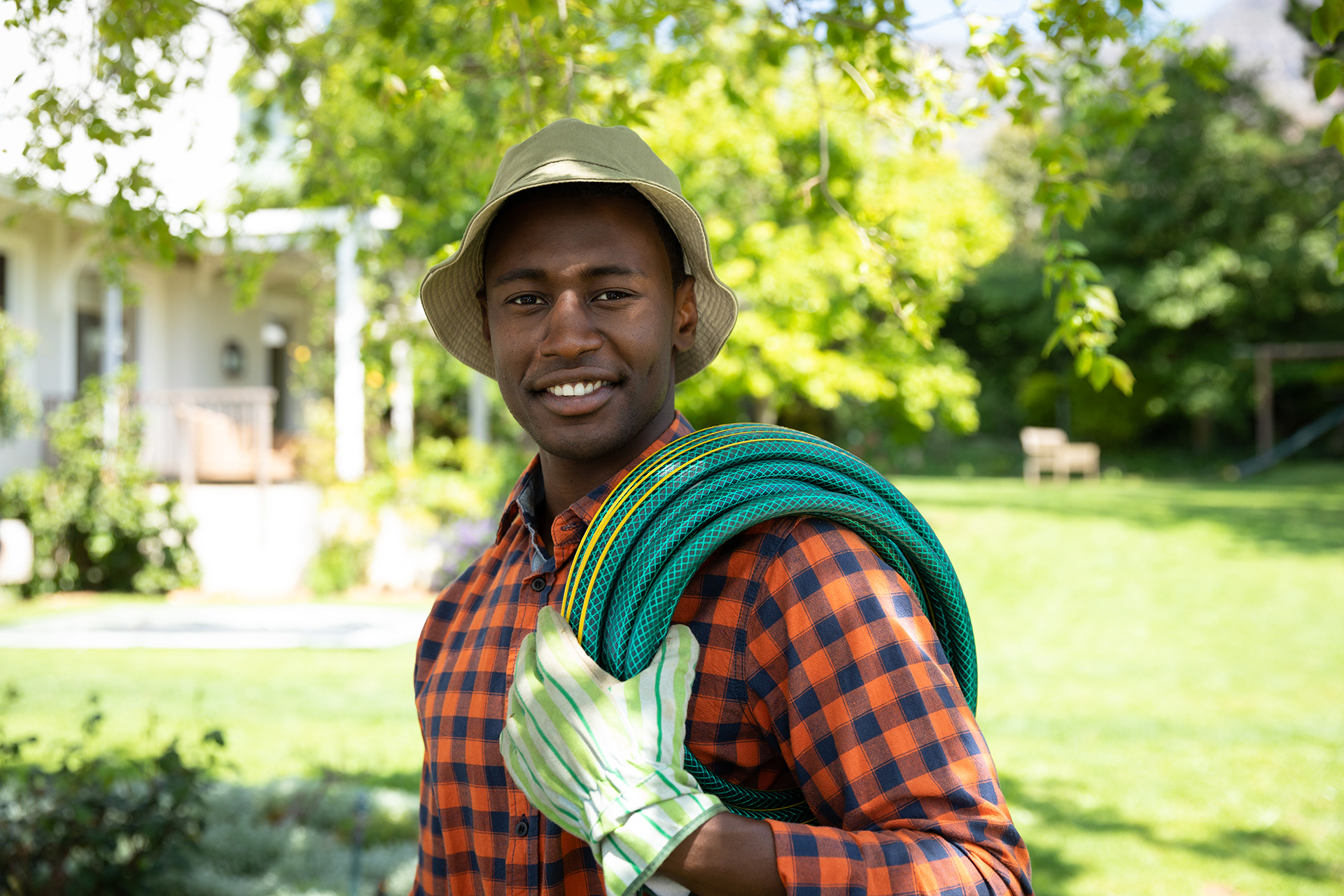 Portrait of an African American man in the garden wearing a hat and gardening gloves, carrying a hose and smiling to camera. Family enjoying time at home, lifestyle concept